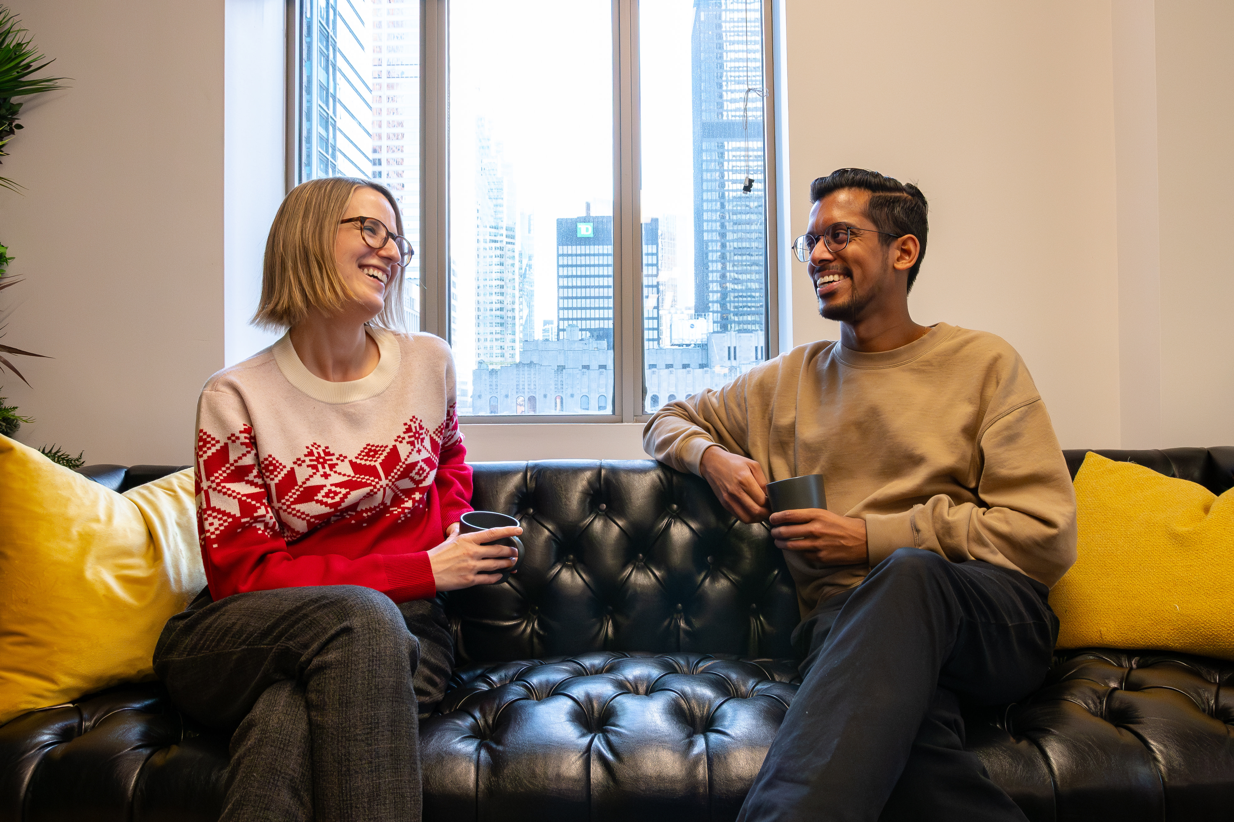 Two professionals sharing a laugh on a leather sofa, coffee in hand, with a city view through the window.