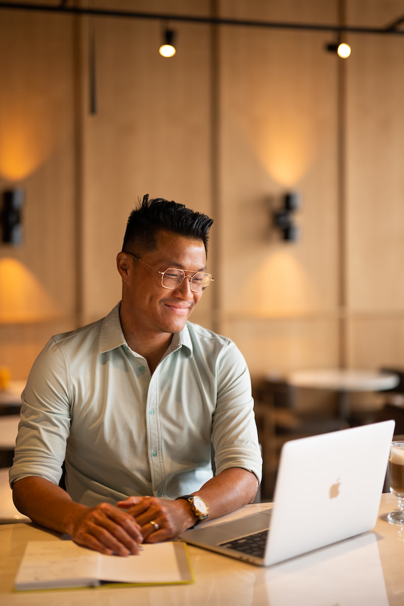 A man wearing a light shirt, with sleeves rolled up, and gold-colored glasses. He is smiling, looking at a laptop screen.