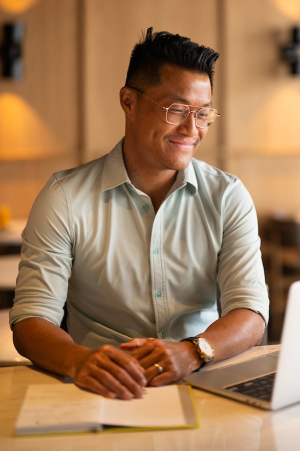 A man wearing a light shirt, with sleeves rolled up, and gold-colored glasses. He is smiling, looking at a laptop screen.