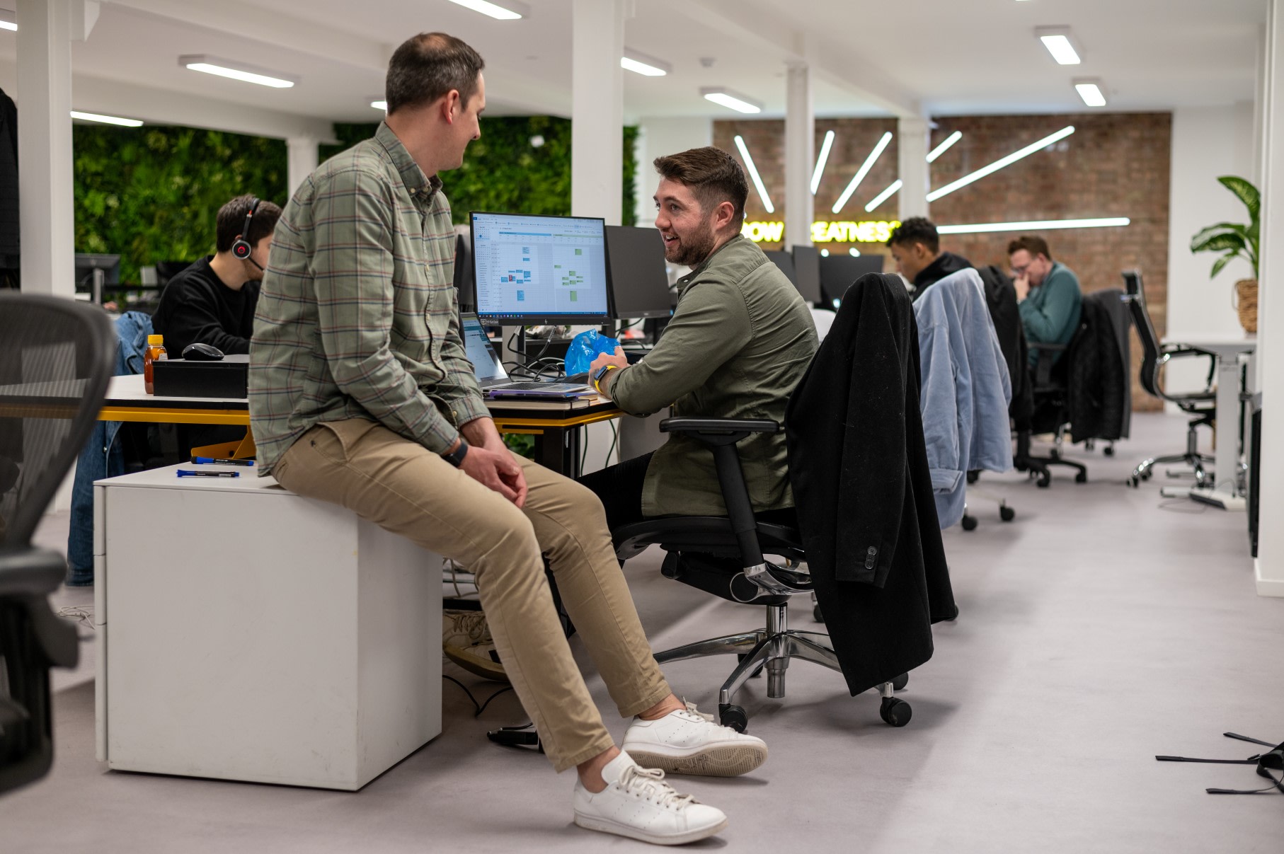 Two men having a discussion in the EZRA offices, one is at a desk, one is sat on a side table