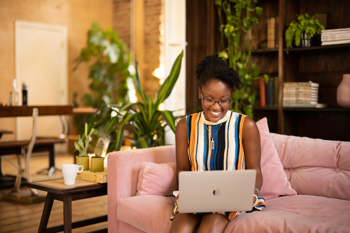 A woman is sat on a pink sofa wearing a colourful top, with her laptop on her lap, smiling at the screen as she takes part in digital coaching.