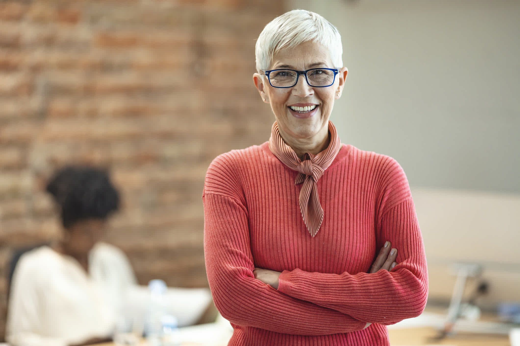 Close-up portrait of mature business woman standing at office. Pretty older business woman, successful confidence with arms crossed in financial building. Mature female in office with team meeting in background.