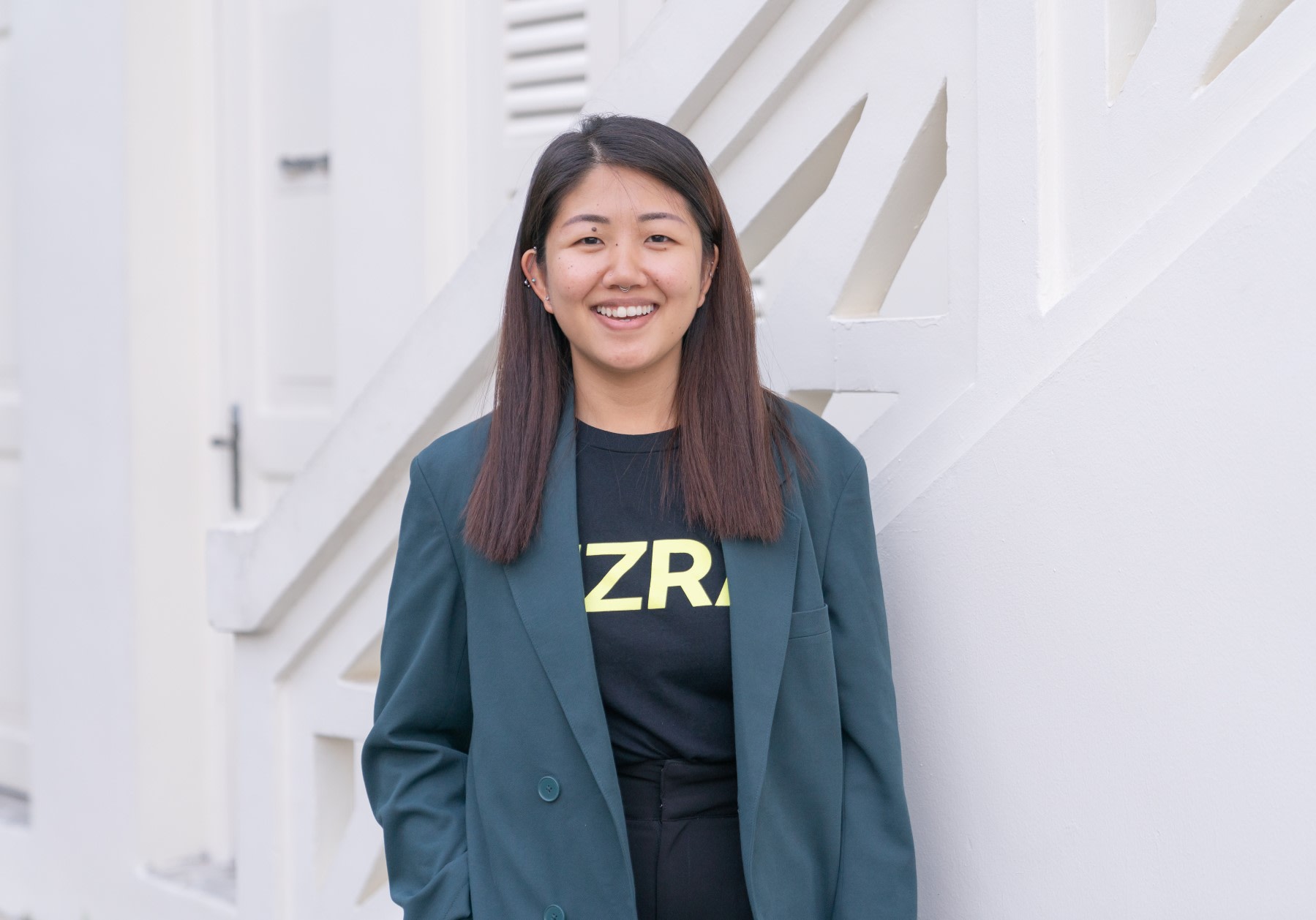 Woman standing in front of a white staircase wearing black trousers, a black top with the EZRA logo printed on the front, and a green blazer on the top.