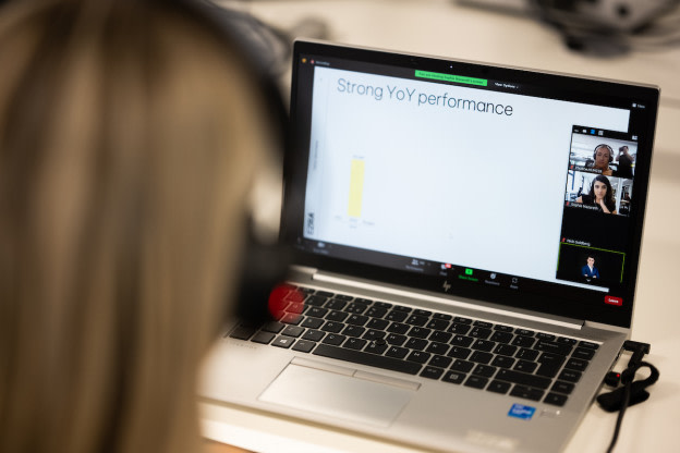 A woman is sat at her laptop with her headphones on, on a call with four other attendees, watching a screen shared presentation on YoY performance.