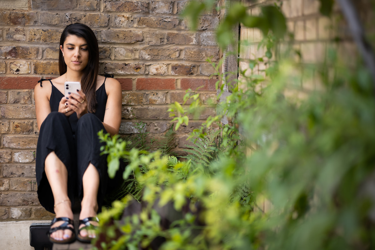 Woman sat with her back to a brick wall, her knees to her chest, on her phone.
