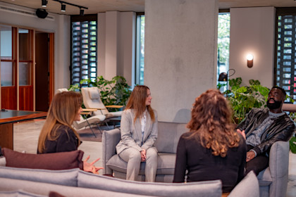 Three women and one man are sat in an office space on sofas having a meeting.