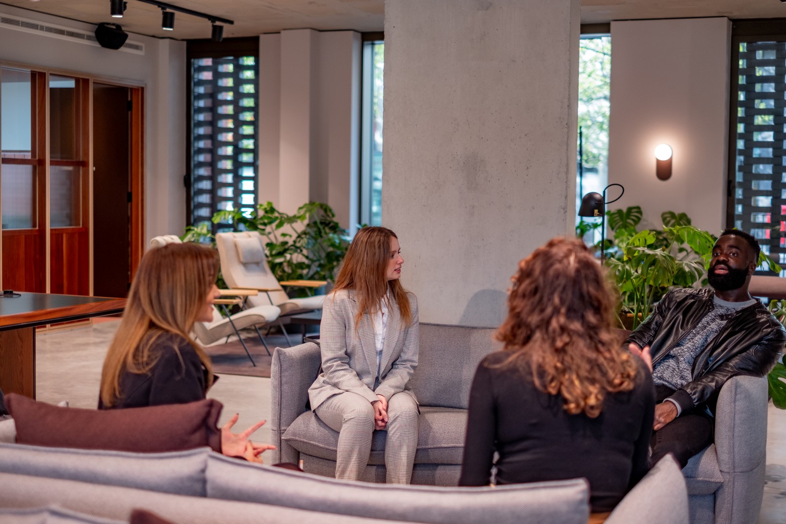 Three women and one man are sat in an office space on sofas having a meeting.