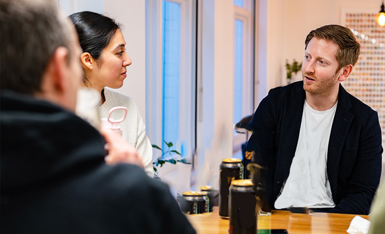 Man wearing dark jacket over white tee shirt, talking about leadership coaching to woman in white top