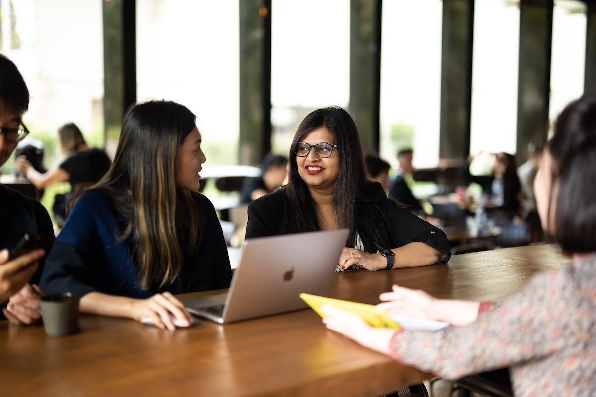 Three women sat at a high table with a laptop and a notepad, discussing things with each other