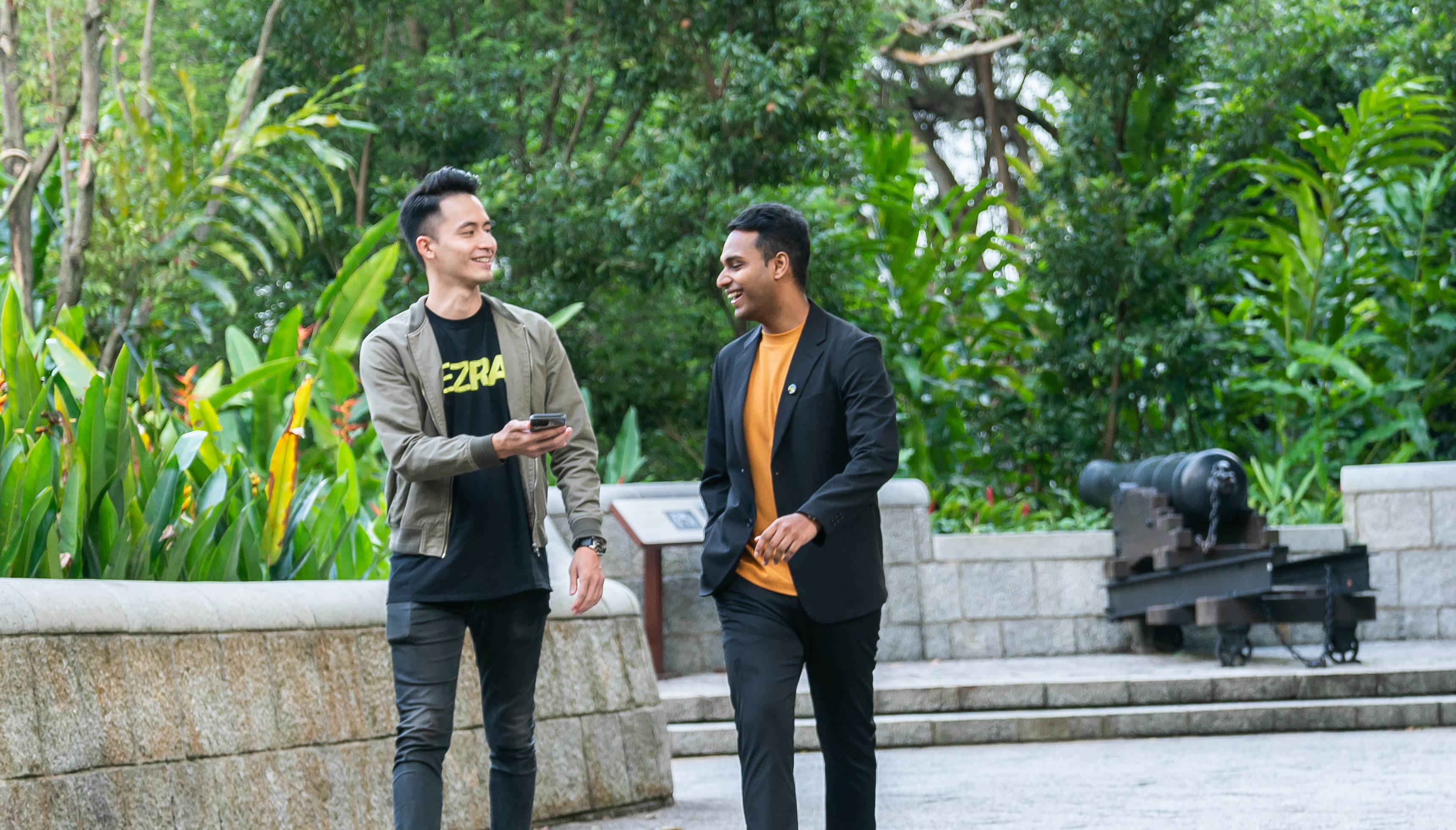 Two smiling men chatting as they walk, one dressed in a grey top over a black EZRA T-shirt, the other in a dark blue jacket over an orange T-shirt