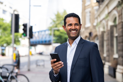 A man stood outside on the sidewalk in a navy suit with his phone in his hand, smiling. 