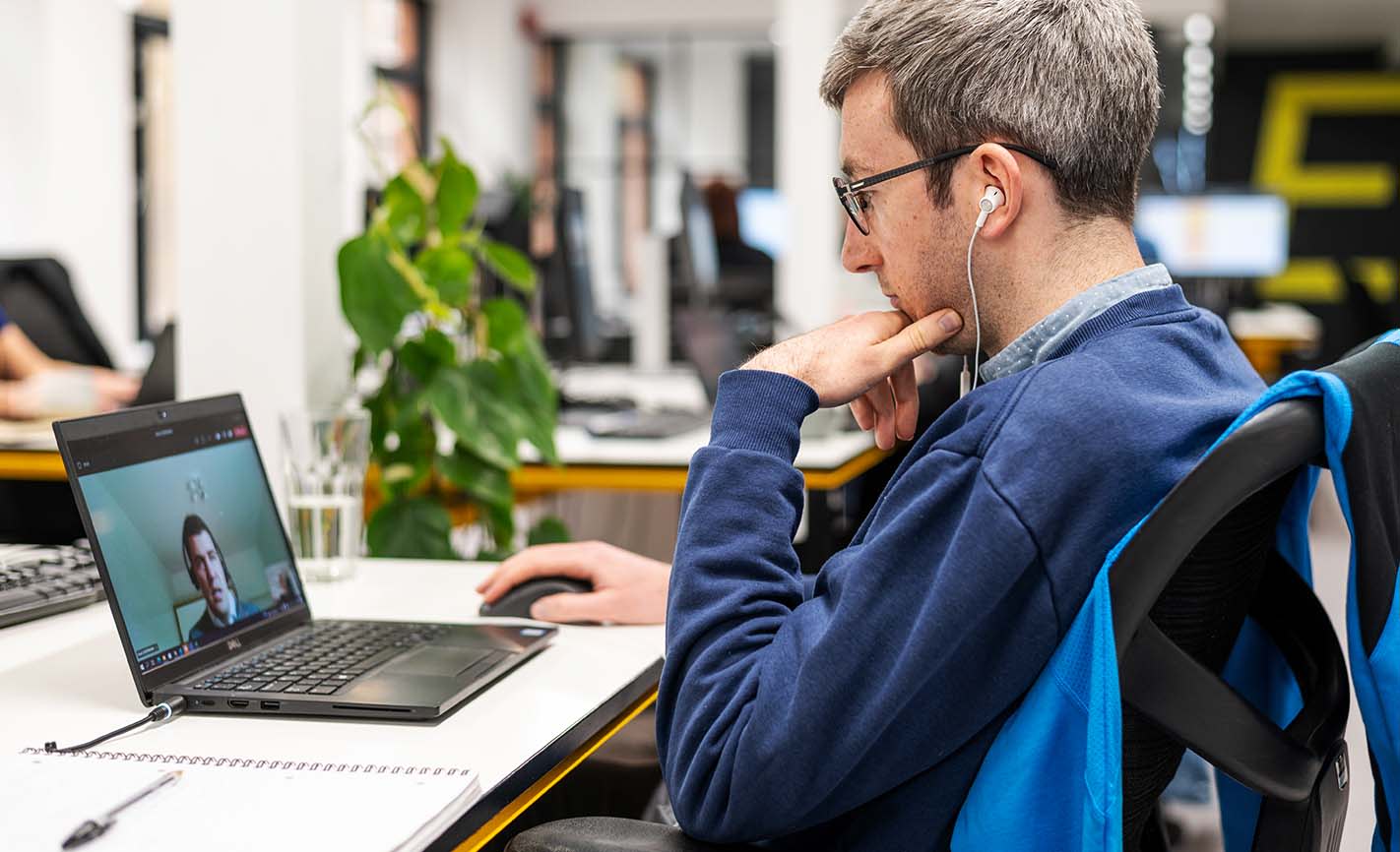 A man in a blue shirt, seated with earbud headphones, watching a virtual coaching session on a laptop