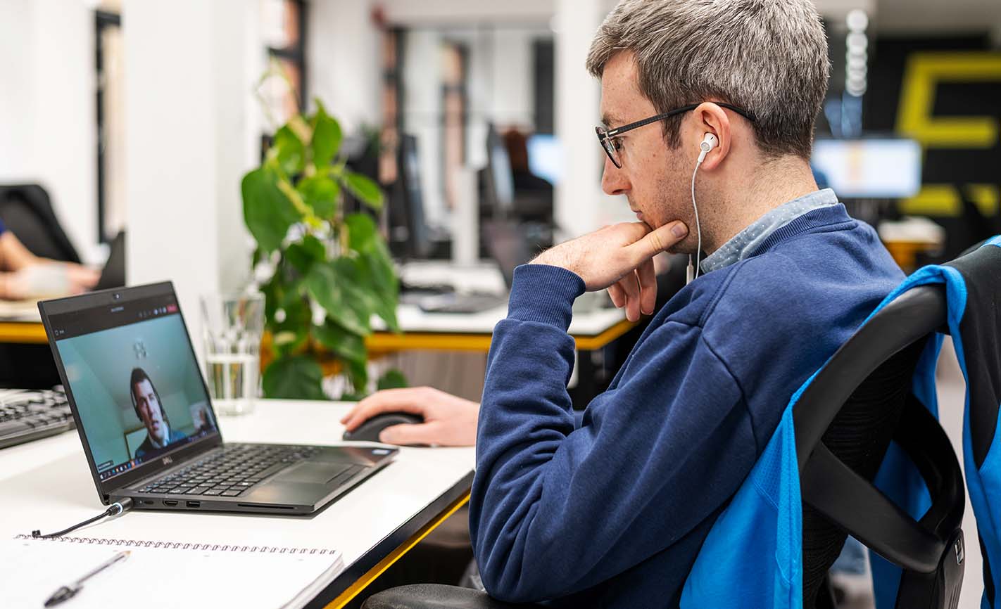 A man in a blue shirt, seated with earbud headphones, watching a virtual coaching session on a laptop
