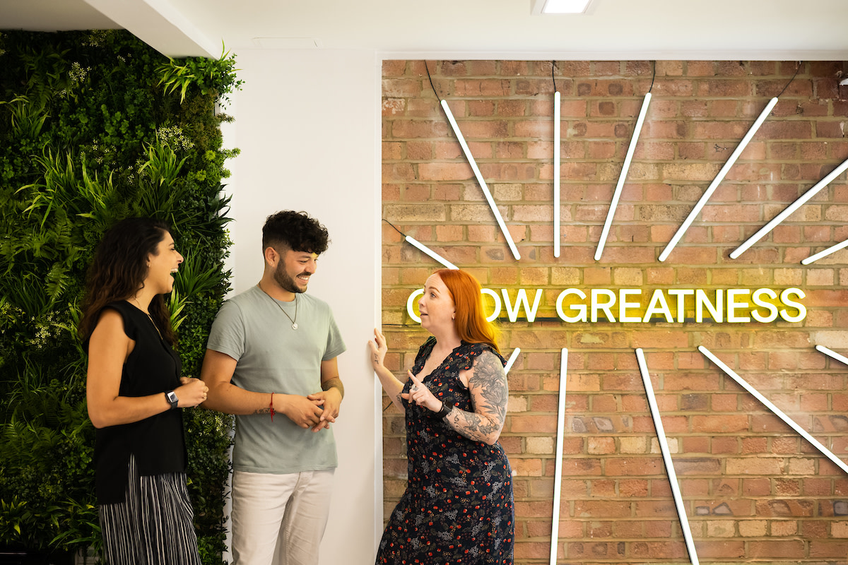 Gruppe von zwei Frauen und einem Mann im Büro von EZRA, die Coaching-Fähigkeiten diskutieren, stehen vor einem gelben Neon-Schild mit der Aufschrift "grow greatness"