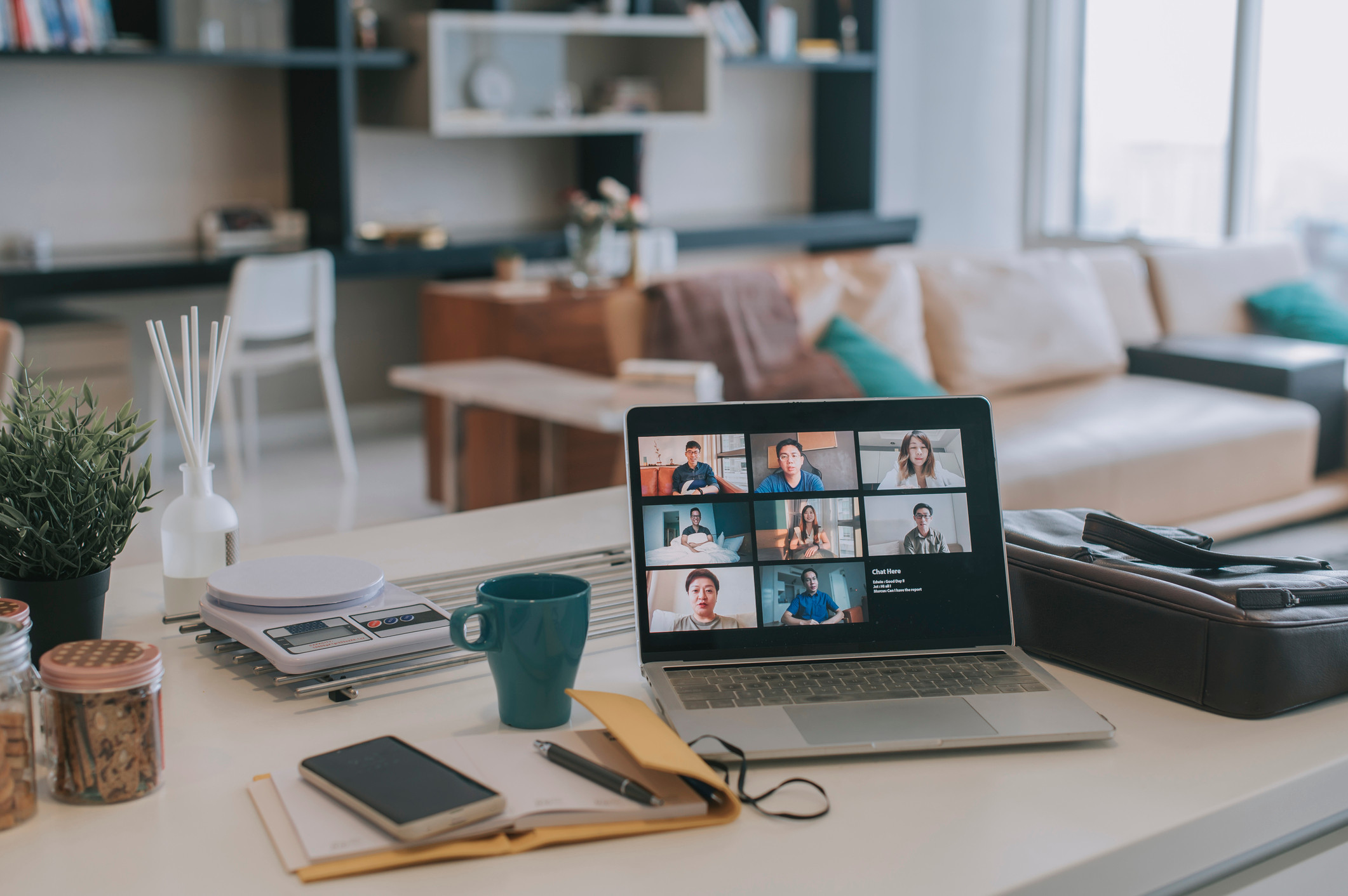 Kitchen counter with a laptop with virtual meeting on it. There is mug, a mobile phone and a notebook next to the laptop, plus various kitchen utensils.