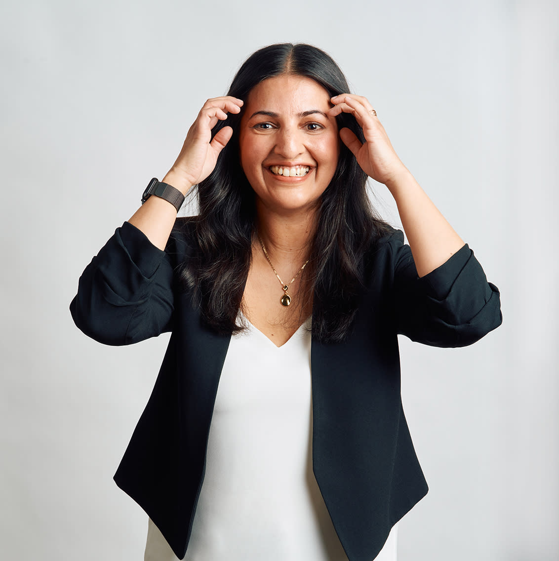 A female manager wearing a white top and a black blazer against a grey background, smiling at the camera and pulling her hair behind her ears.