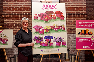 Anne Geddes stands next to a large print of artwork on an easel featuring babies in pink pots next to Wave petunias.