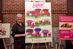 Anne Geddes stands next to a large print of artwork on an easel featuring babies in pink pots next to Wave petunias.