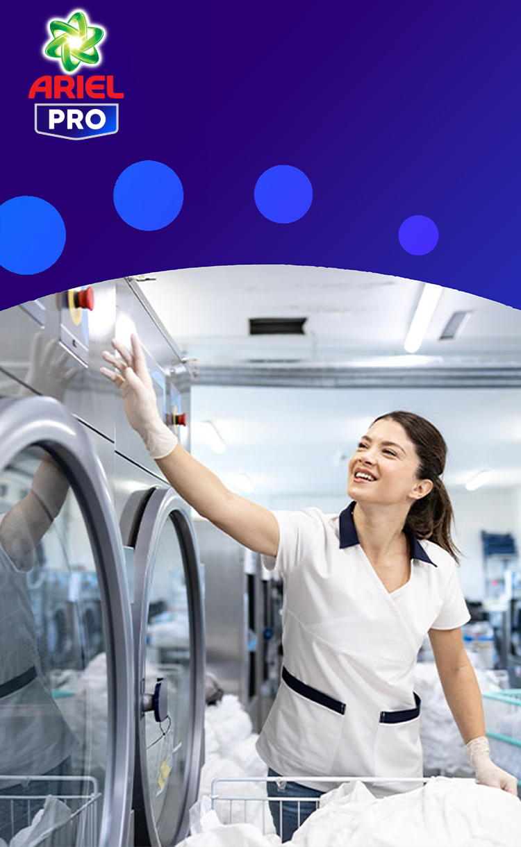 Mujer sonriente con uniforme usa una lavadora industrial en una lavandería profesional. Logo de Ariel PRO sobre un fondo azul.
