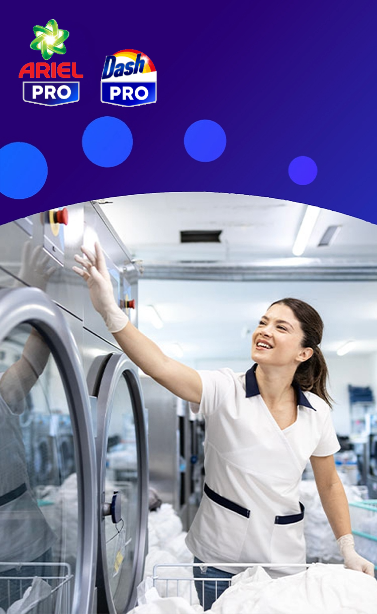 Een lachende vrouw in een wit uniform en handschoenen werkt in een professionele wasserette en reikt naar een rij grote, roestvrijstalen industriële wasmachines. De logo's van Ariel Pro en Dash Pro staan links op een paarse achtergrond.