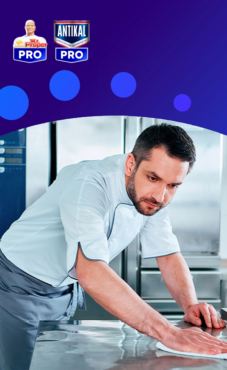 Un chef nettoyant un comptoir en acier inoxydable dans une cuisine professionnelle, avec les logos Mr. Propre Pro et Antikal Pro.