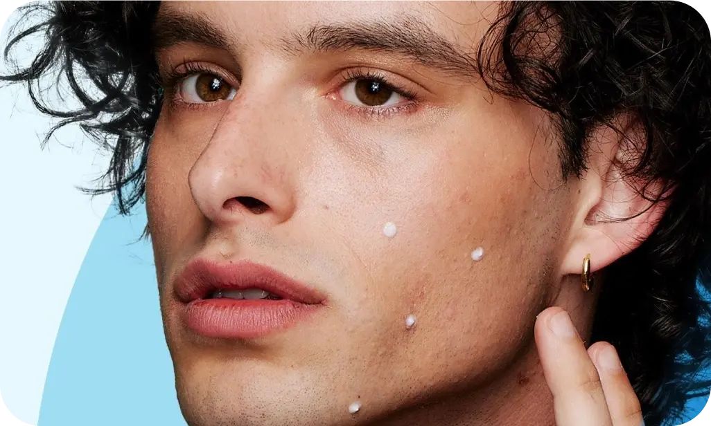 A person with curly hair applies small white dots of cream on their face, focusing intently. The background features light blue and darker blue curves.