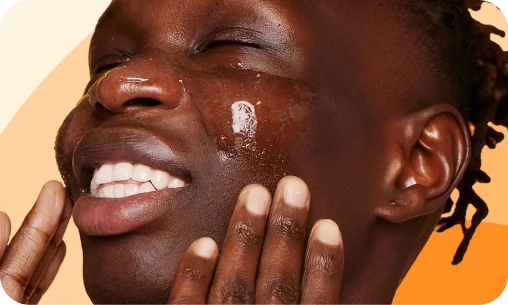 A person smiles with eyes closed, applying a glossy skincare product on their face, using their hands. The background features a gradient of light to dark orange hues.