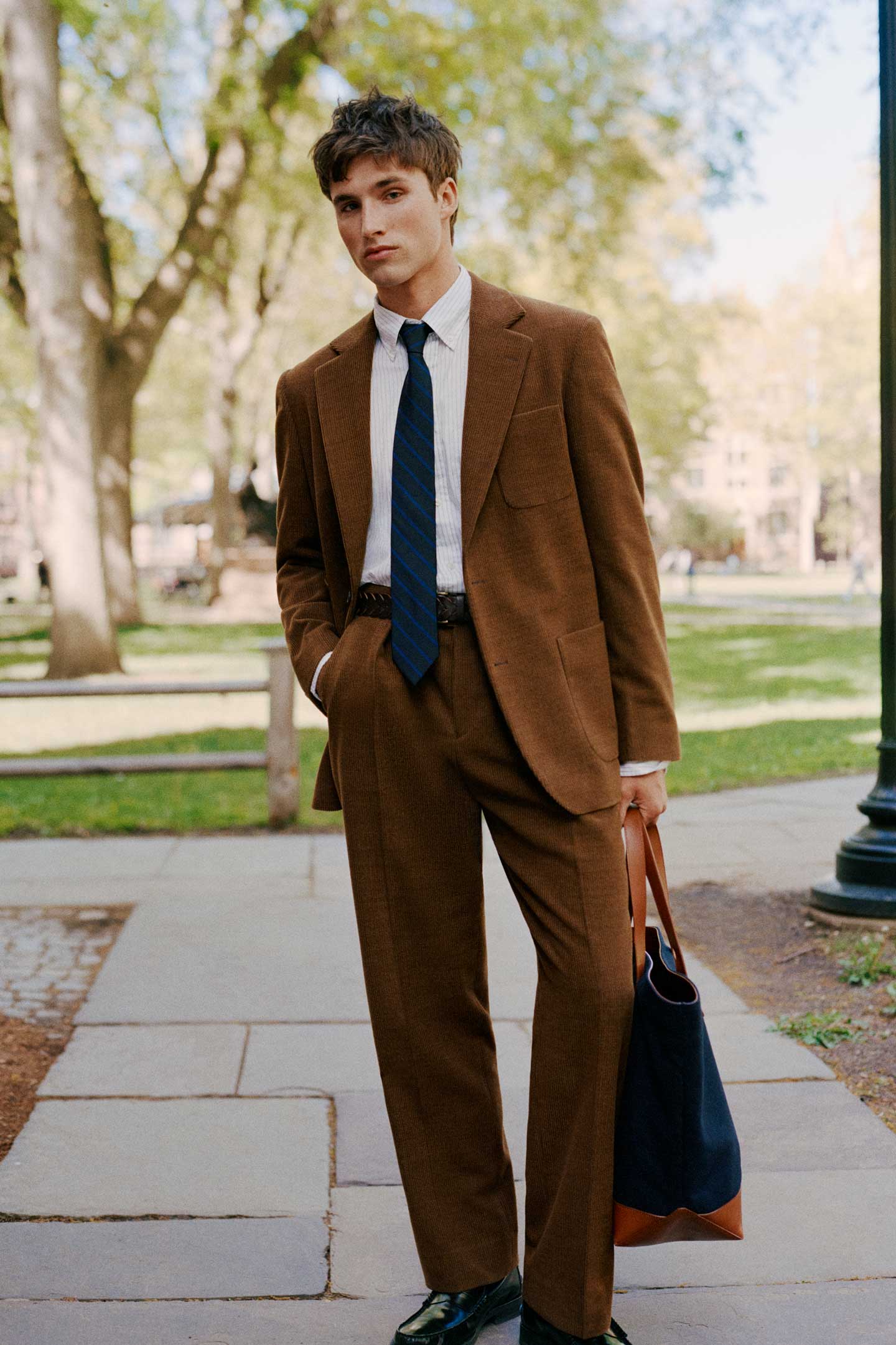 Model styled in a GANT brown suit with shirt and tie, standing outdoors in a campus setting.