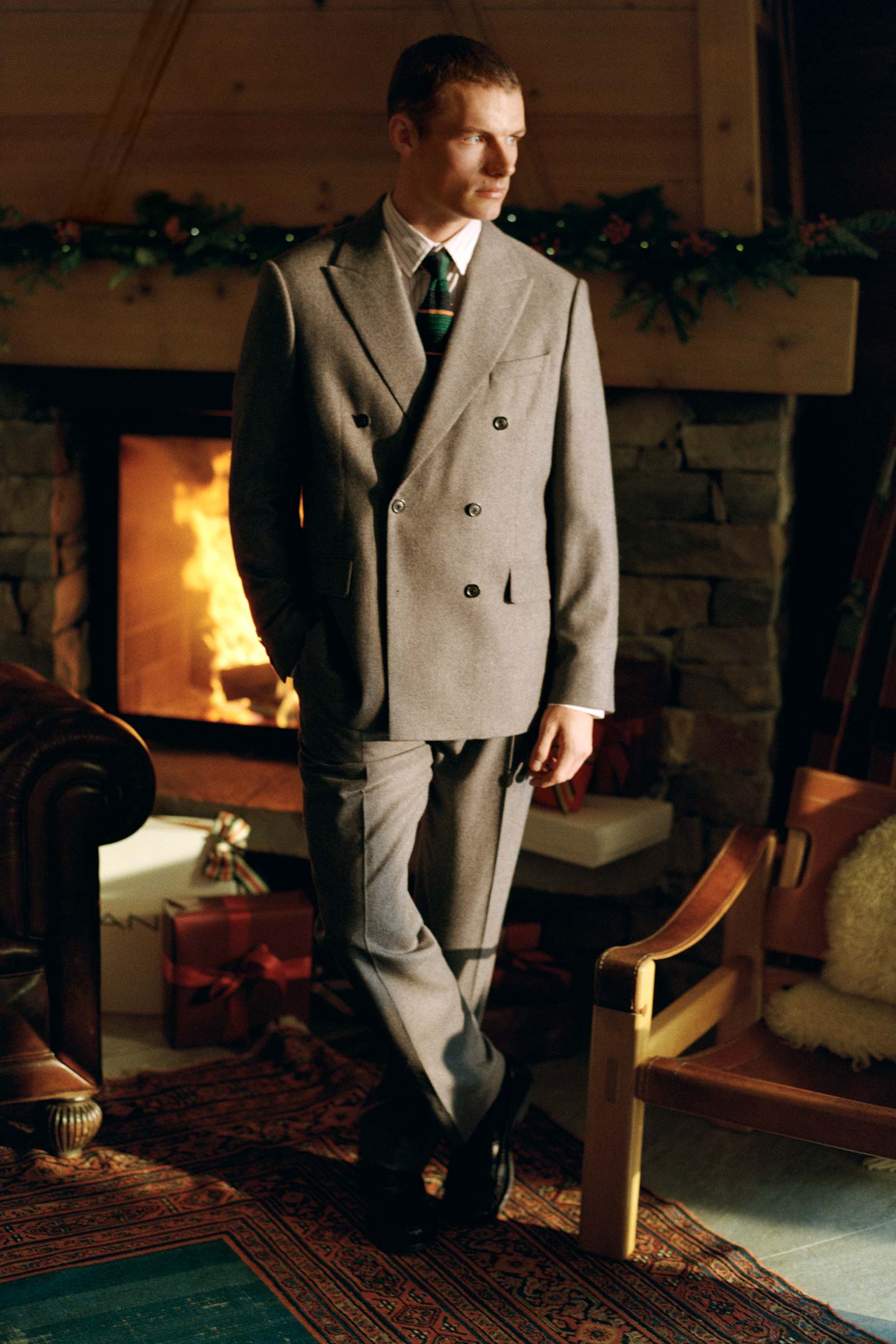 Model styled in a Gant double-breasted suit worn with a shirt and striped tie beside a fireplace.