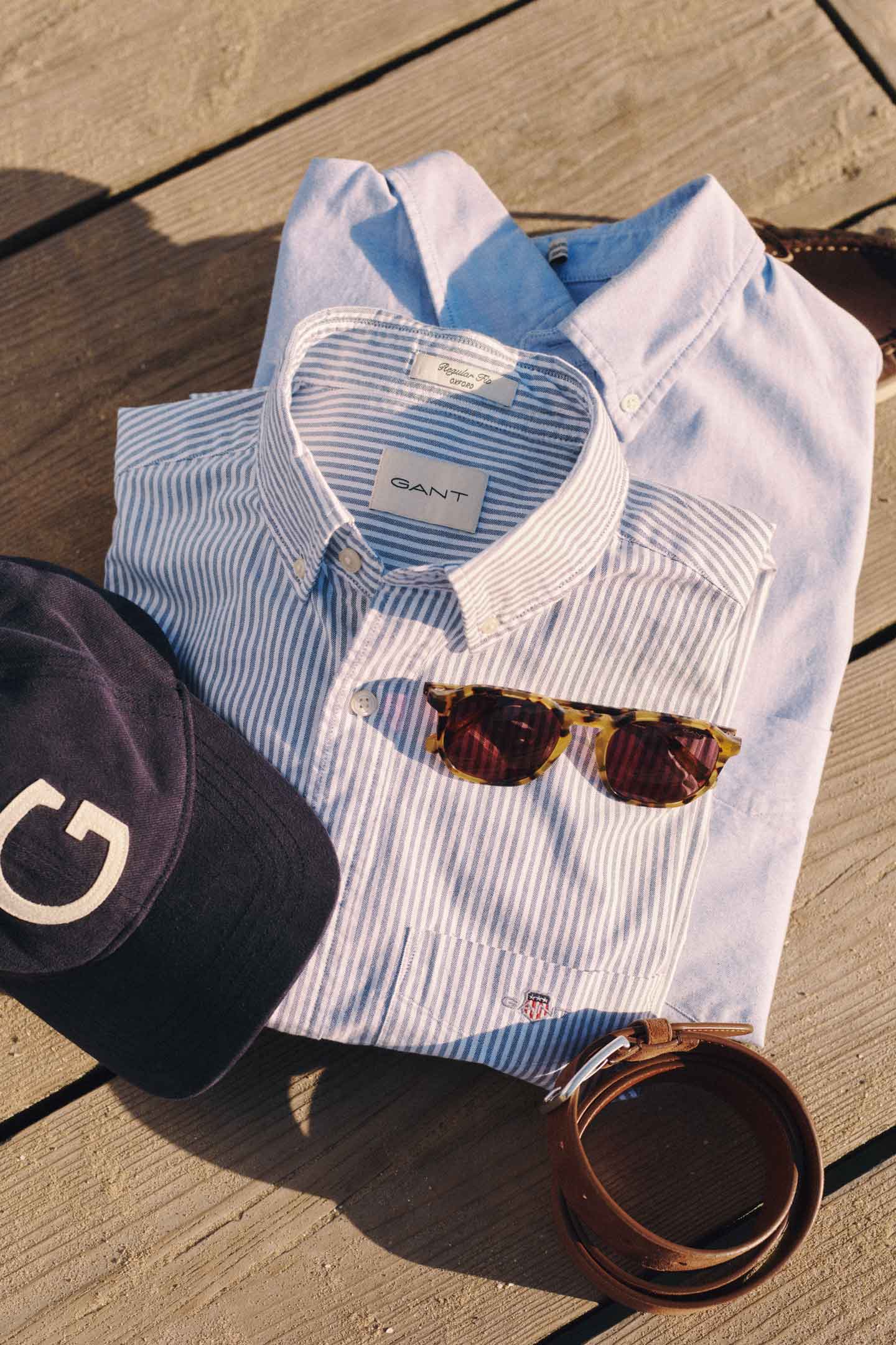 Gant striped and solid button‑down shirts styled with a navy cap, tortoiseshell sunglasses, and a brown leather belt on a wooden surface.