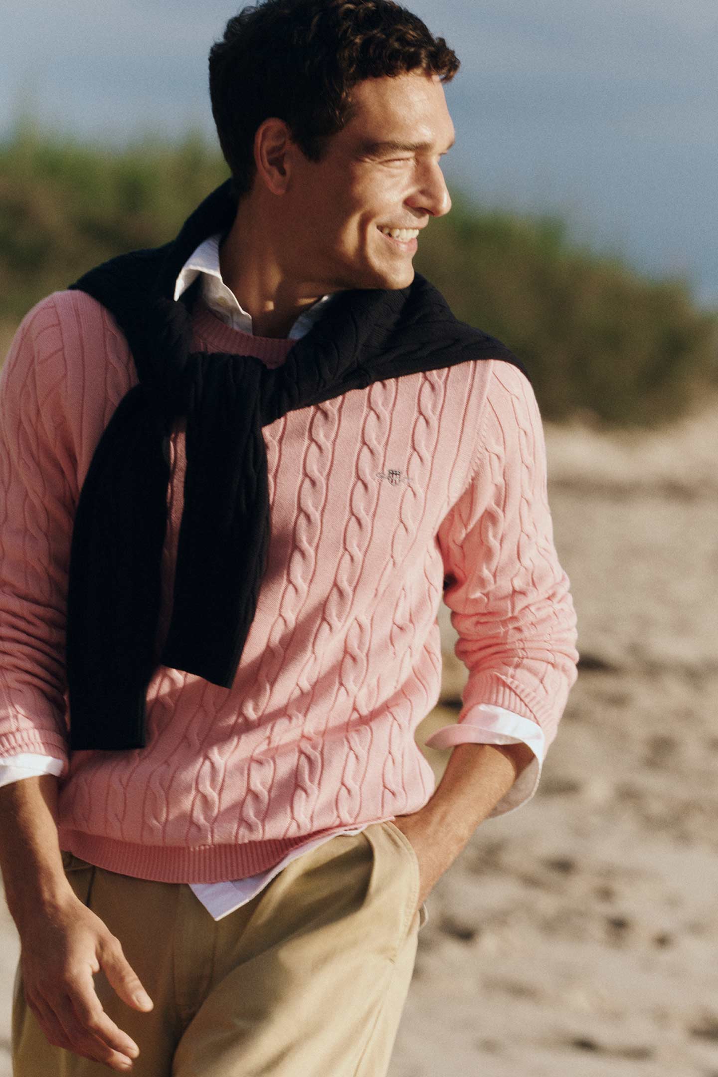 Men wearing Gant pink cable knit sweater styles with a black sweater over the shoulders and beige trousers on a beach.