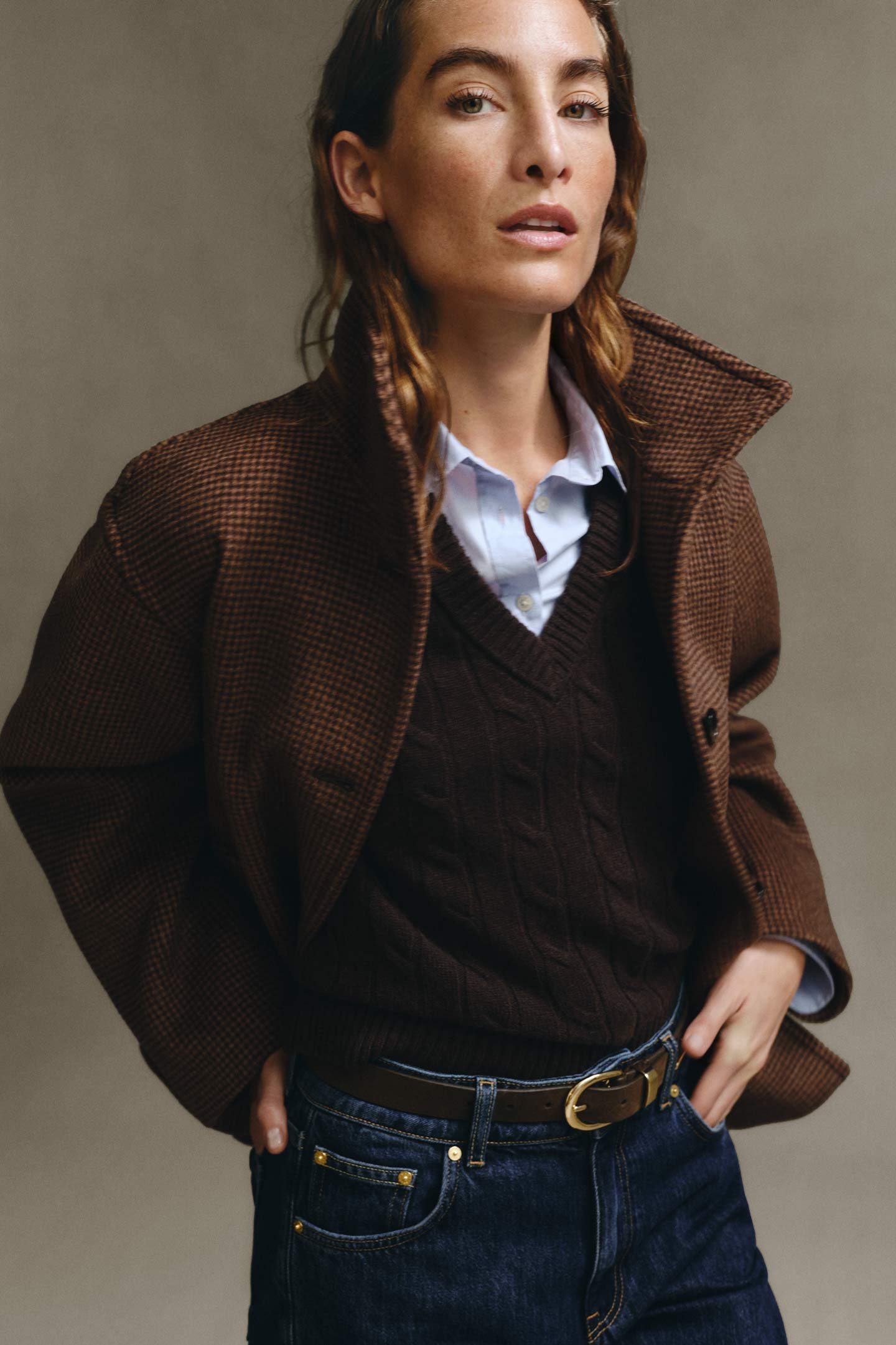 Model wearing a brown textured blazer layered over a dark brown cable-knit sweater and light blue button-down shirt, paired with dark wash jeans and a leather belt.