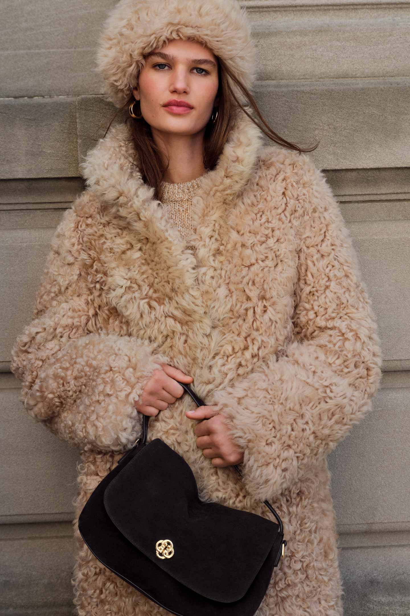 Model styled in a Gant shearling coat and matching hat and a knit sweater, holding a black shoulder bag.