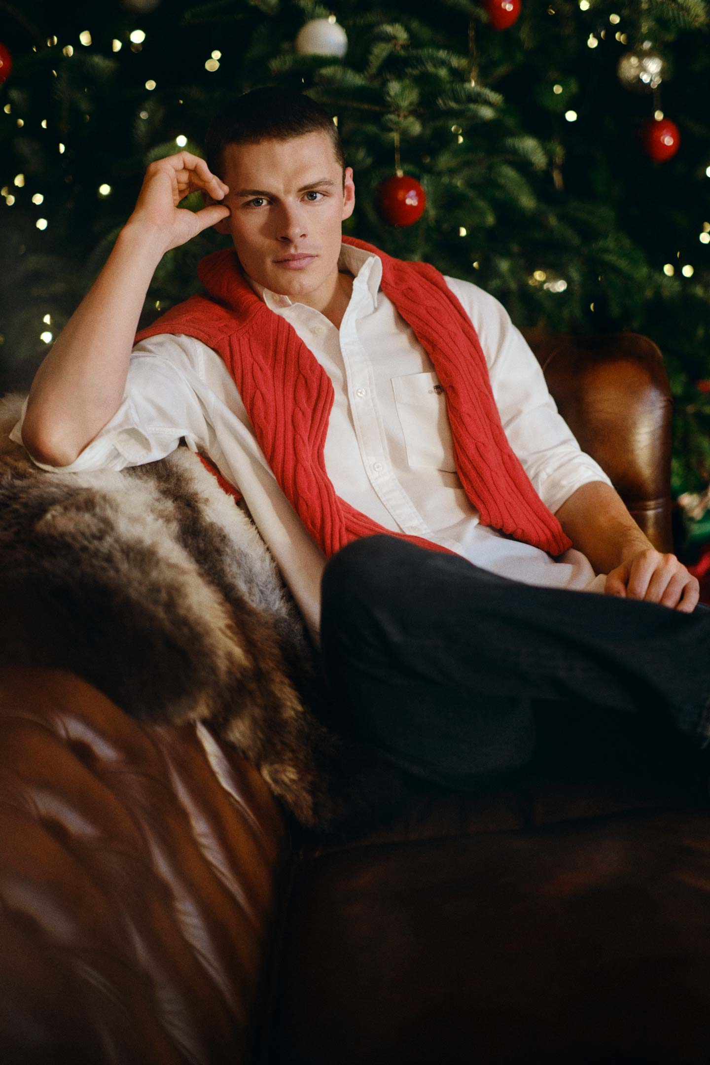 Model styled in a Gant button-down shirt paired with a cable-knit sweater draped over the shoulders, seated indoors.