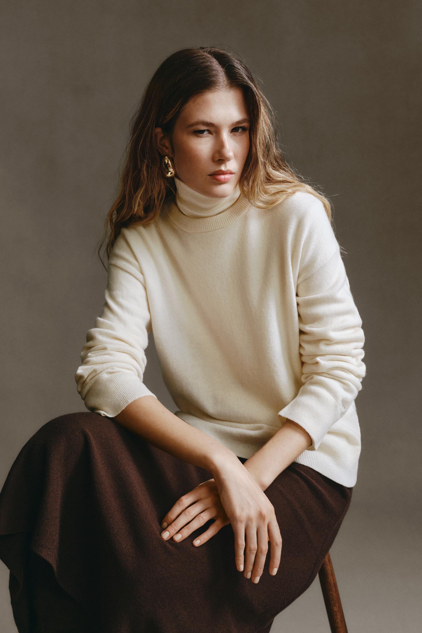 Model styled in a Gant roll-neck sweater paired with a midi skirt, seated on a wooden stool in a studio setting.