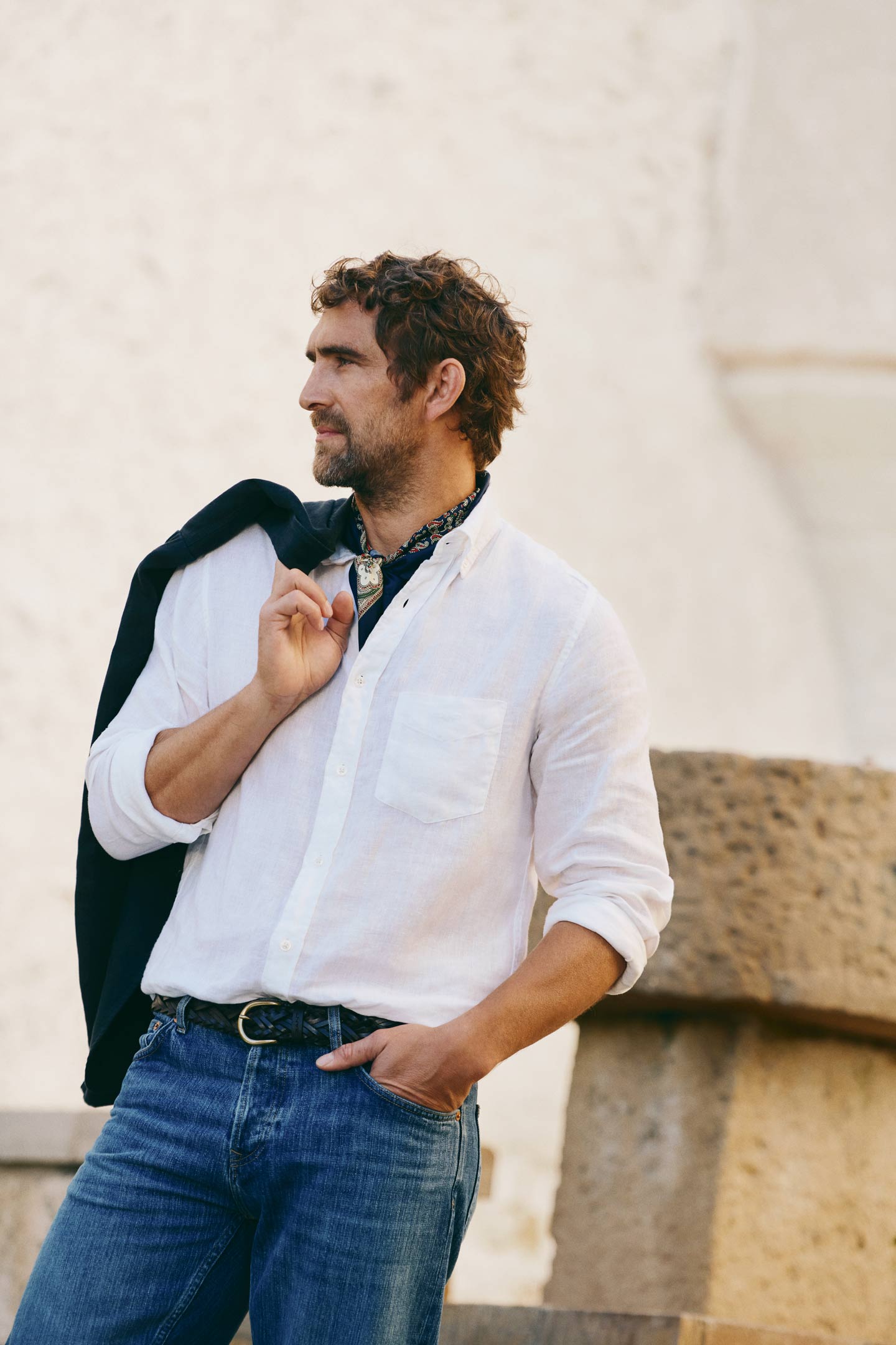 Man wearing a Gant white linen shirt with jeans, belt and a blazer hanging over shoulder in an outdoor setting.