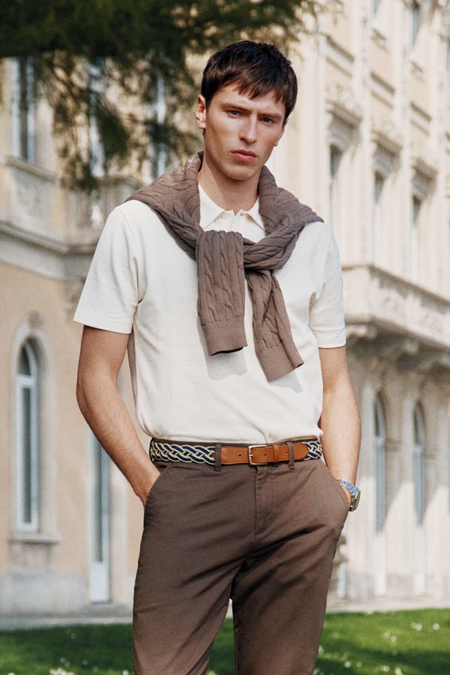 Model styled in a GANT cream polo shirt and tailored brown trousers standing outdoors near a classic building.