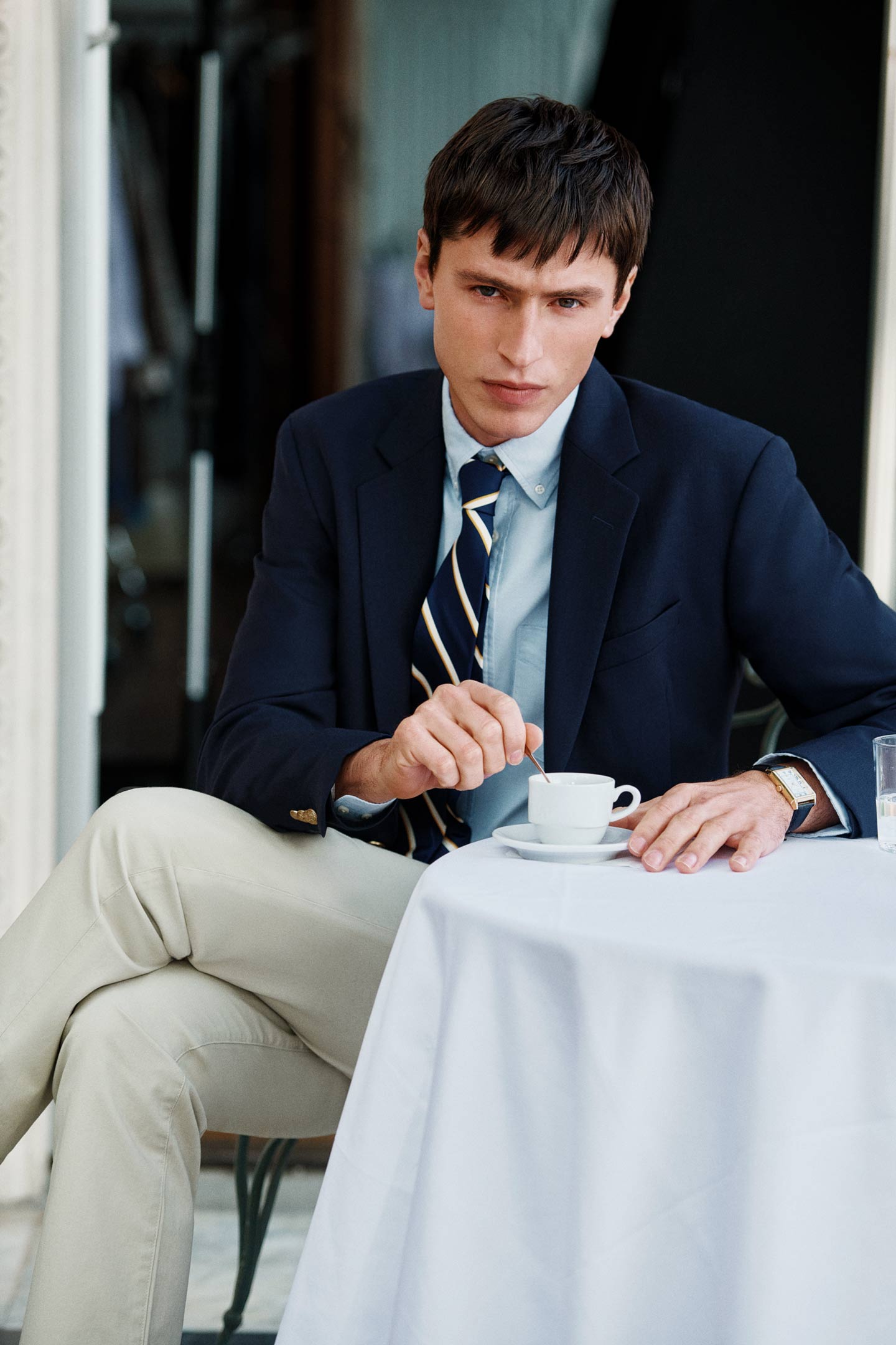 Model styled in a GANT navy blazer with a shirt, tie and light trousers sitting at an outdoor table.