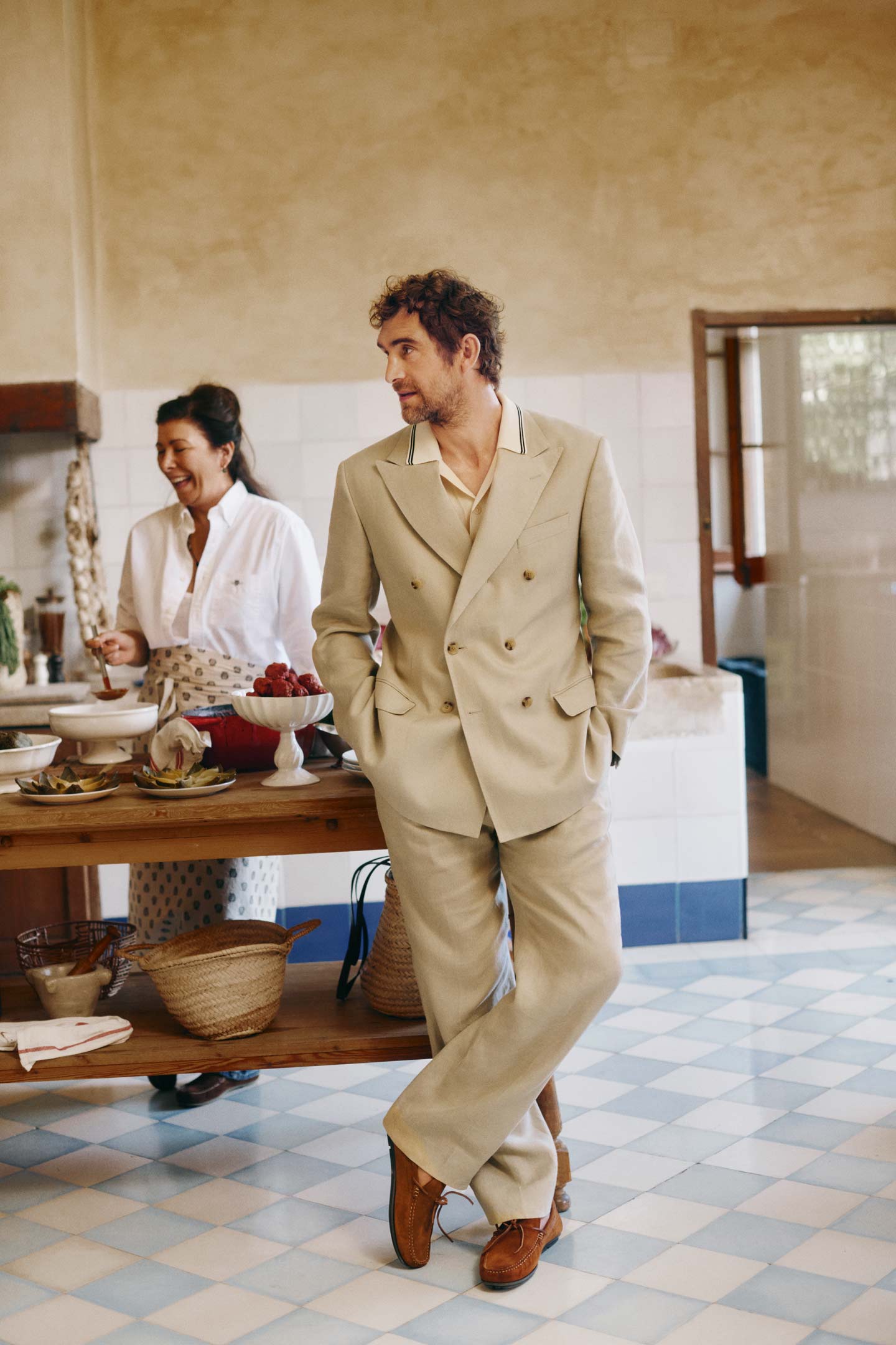 Man wearing a Gant beige double‑breasted suit standing in a kitchen beside someone preparing food