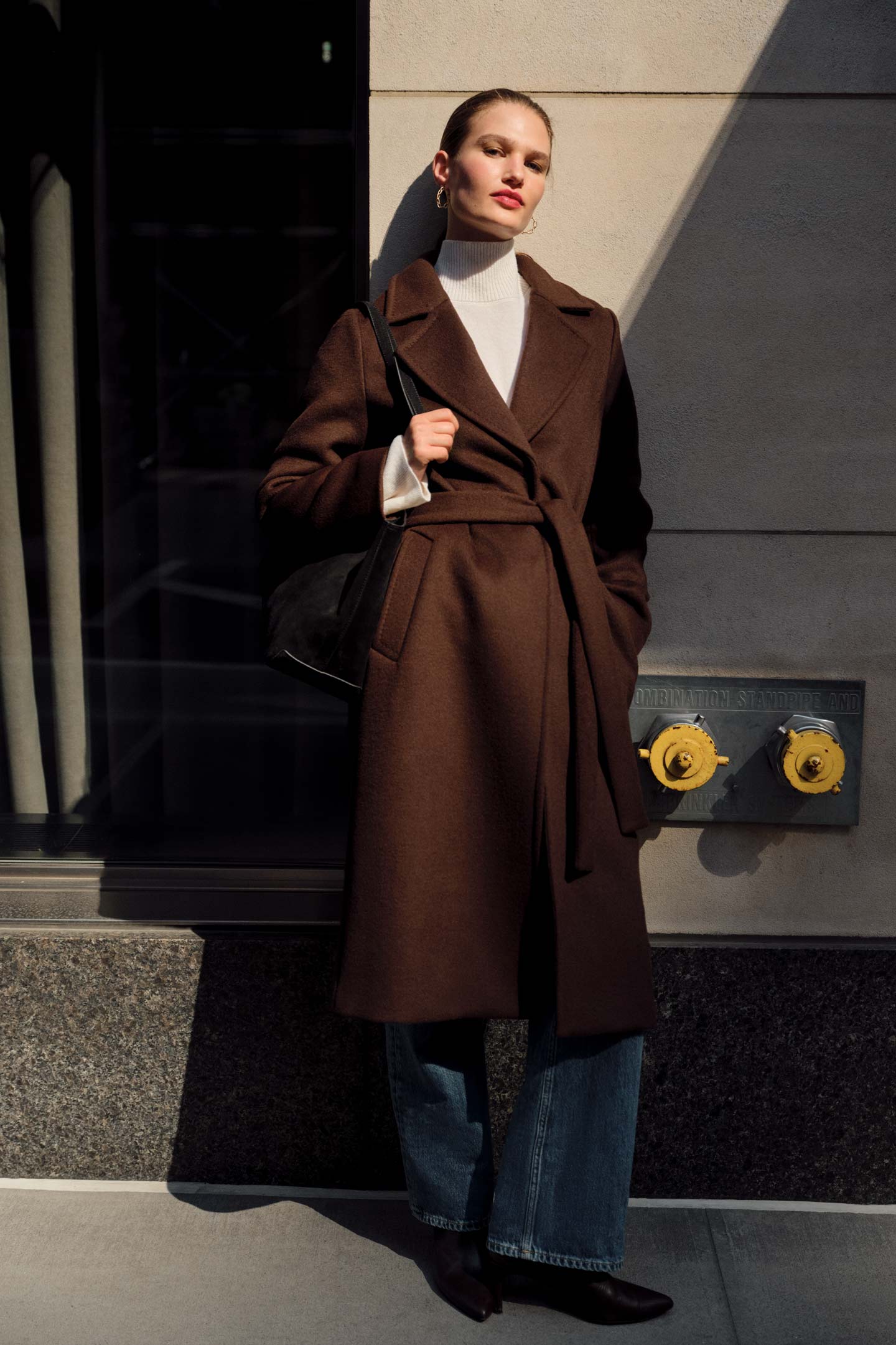 Model styled in a Gant belted wool coat styled with a roll-neck sweater and wide-leg jeans, leaning against a building wall.