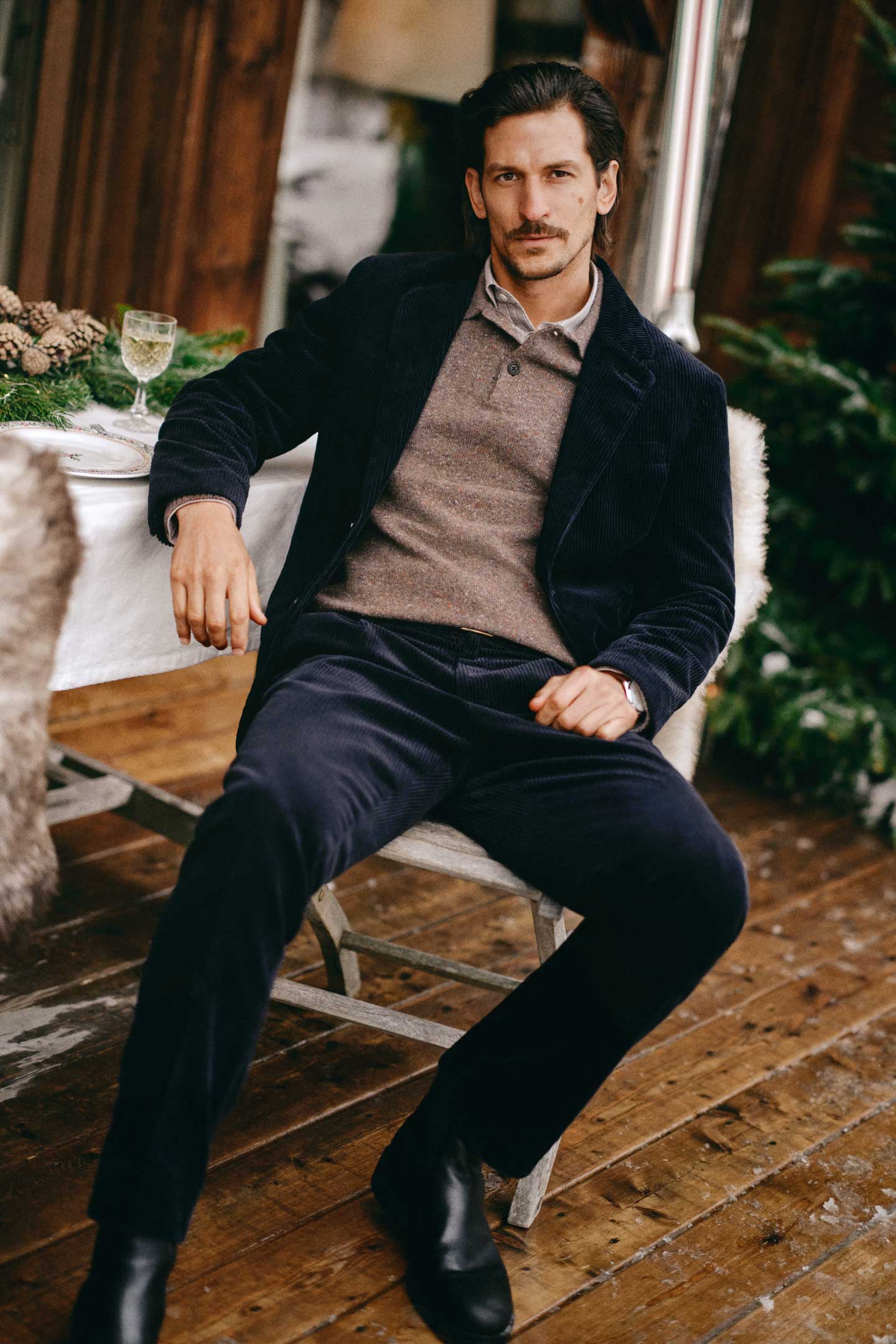 Model styled in a Gant corduroy suit layered over a knit polo sweater, seated at an outdoor dining table.
