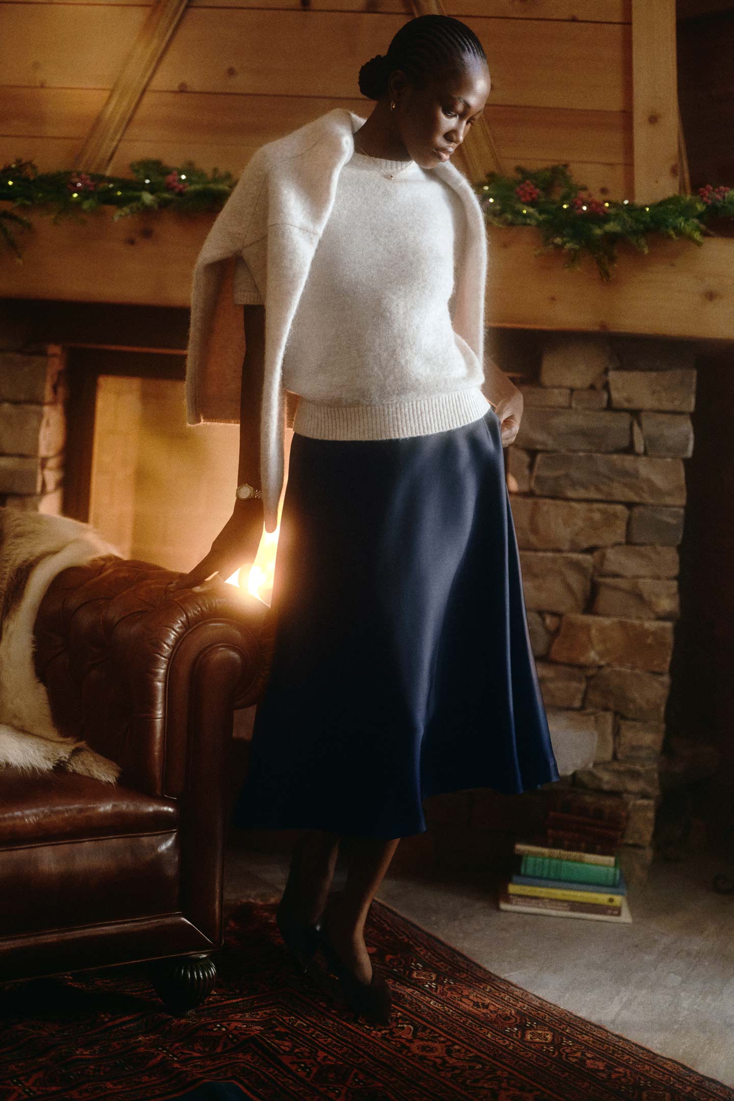 Model styled in a Gant knit sweater draped over the shoulders and paired with a satin midi skirt beside a fireplace.
