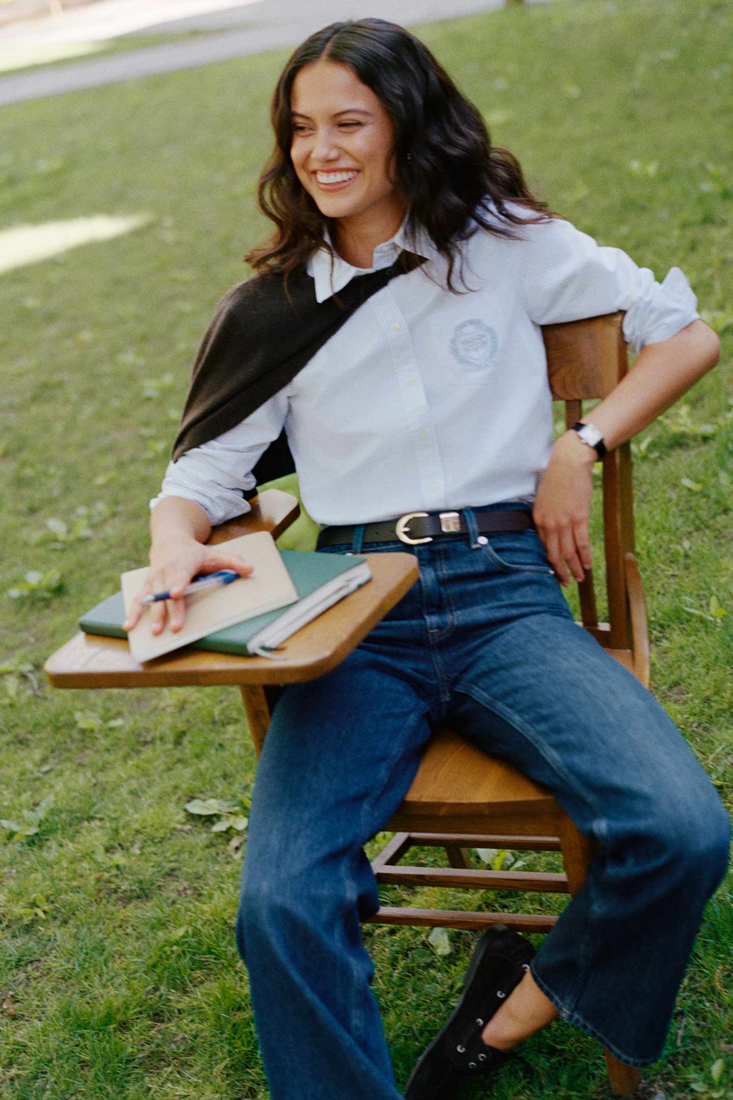Model styled in a GANT white shirt with logo, dark jeans and brown cardigan over shoulder, sitting on a chair in an outdoor campus setting.