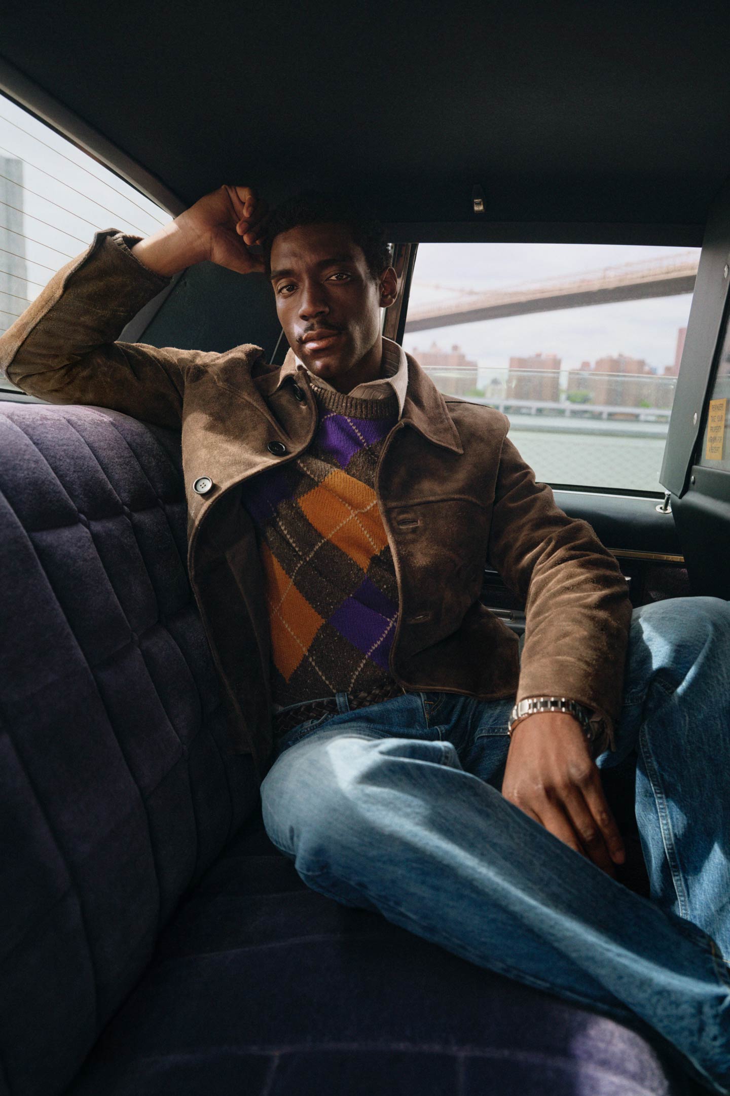 Model styled in a Gant suede jacket layered over an argyle knit sweater and jeans, sitting in the back of a yellow taxi.