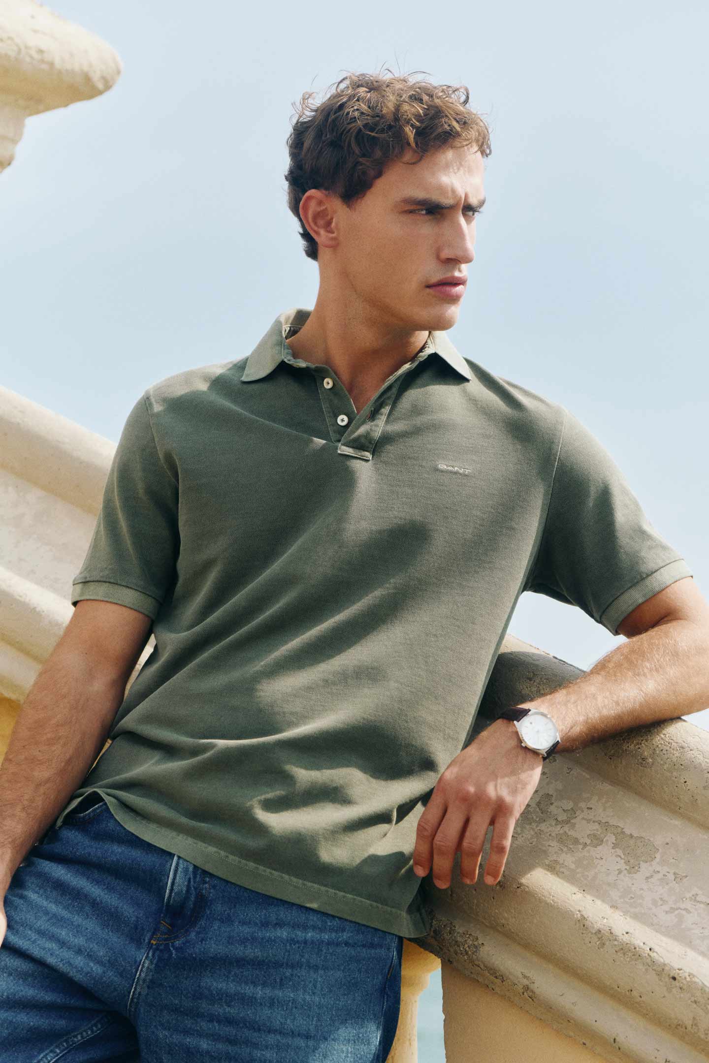 Man wearing a Gant green polo shirt with denim jeans leaning on a stone railing outdoors.