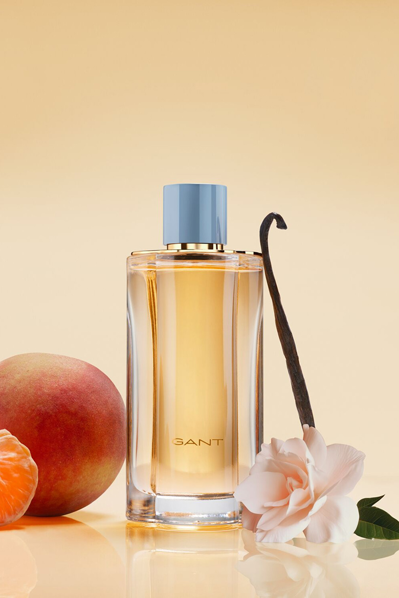 Flacon de parfum GANT pour femme aux tons ambrés entouré d’une pêche, de tranches d’orange, de vanille et d’une fleur rose pâle sur fond clair