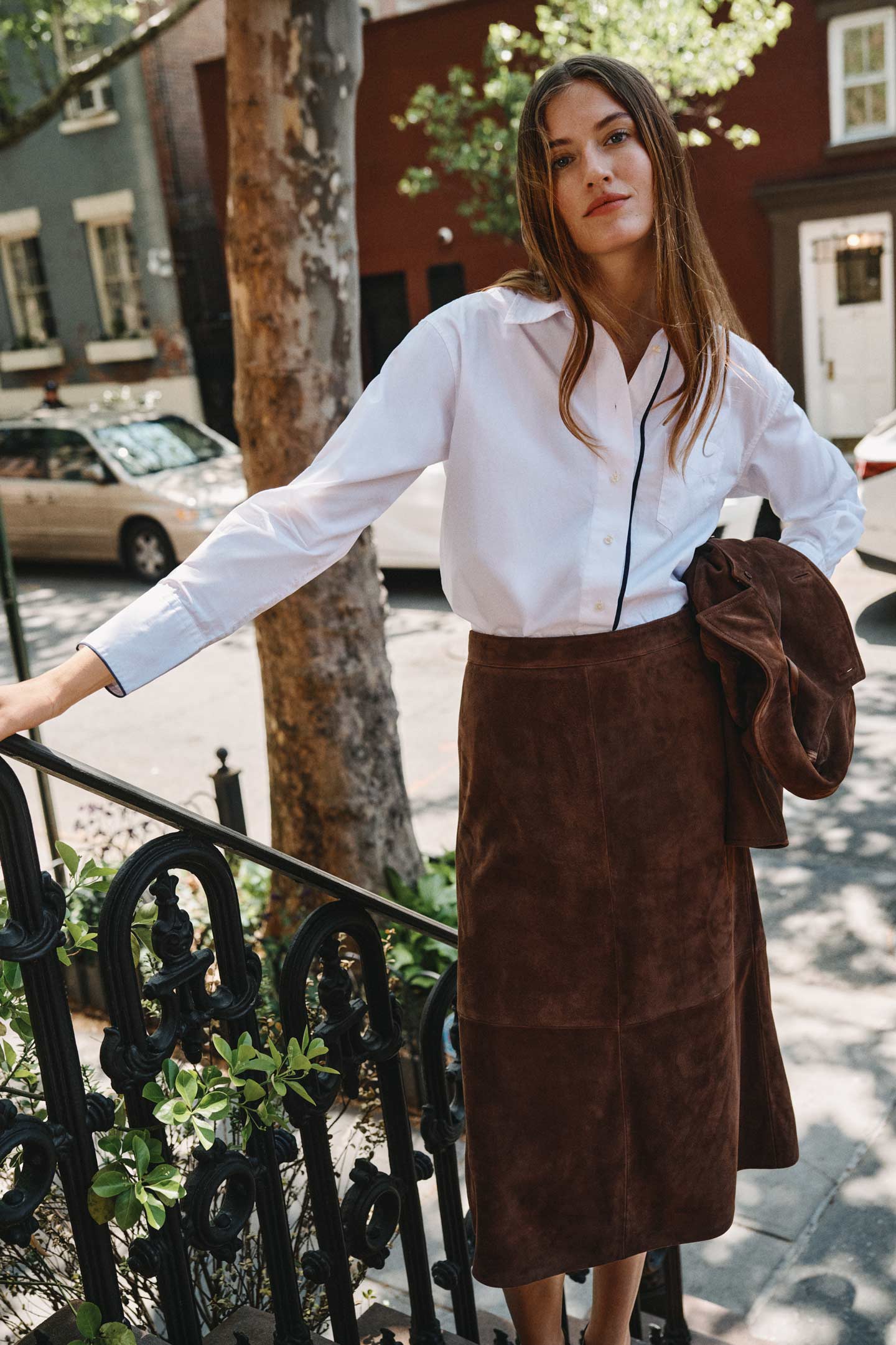 Model styled in a Gant white shirt and suede skirt, holding a suede jacket and standing on steps outdoors.
