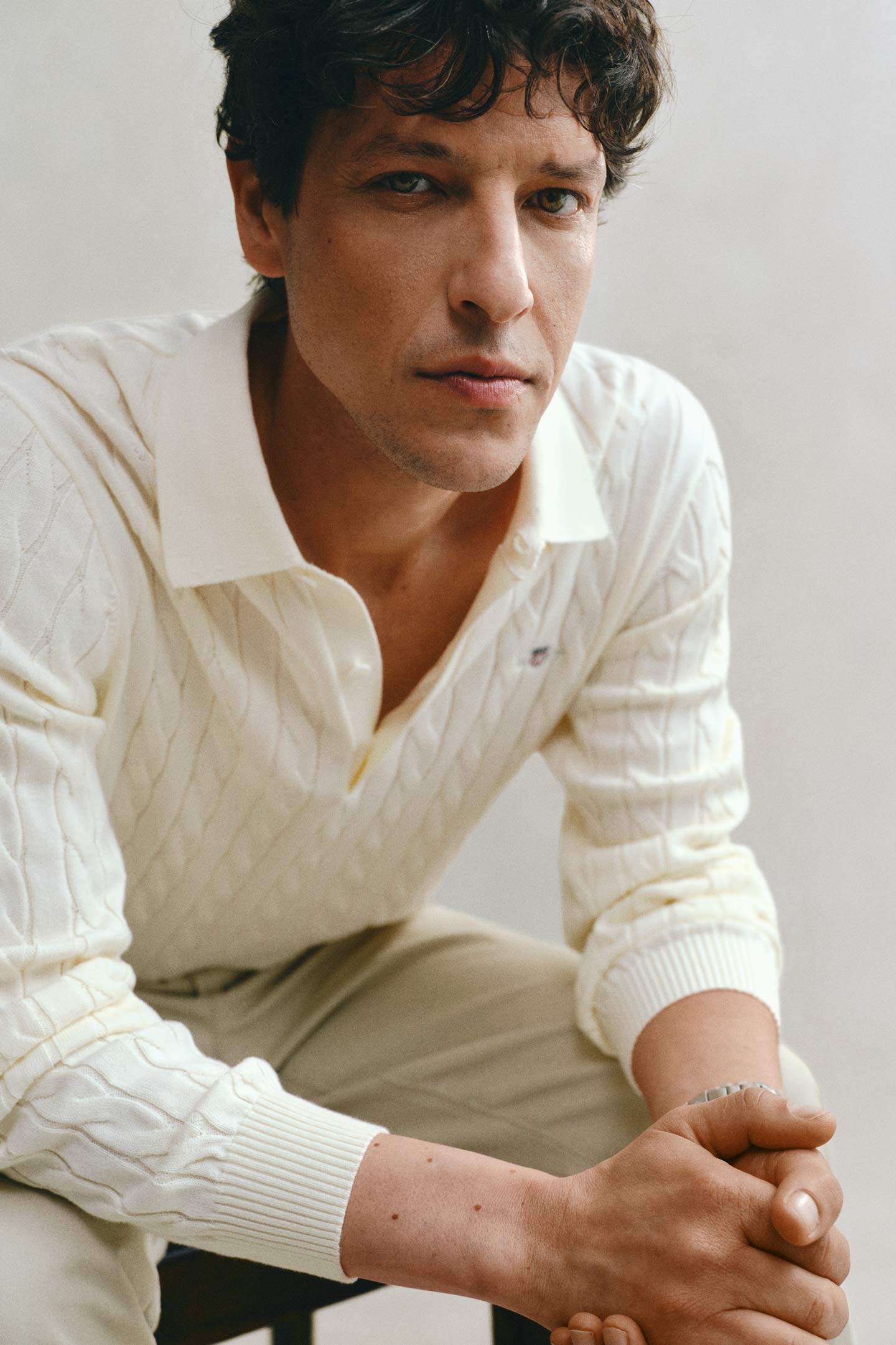 Model styled in a GANT cream cable knit rugger paired with tailored pants sitting in front of a grey backdrop.