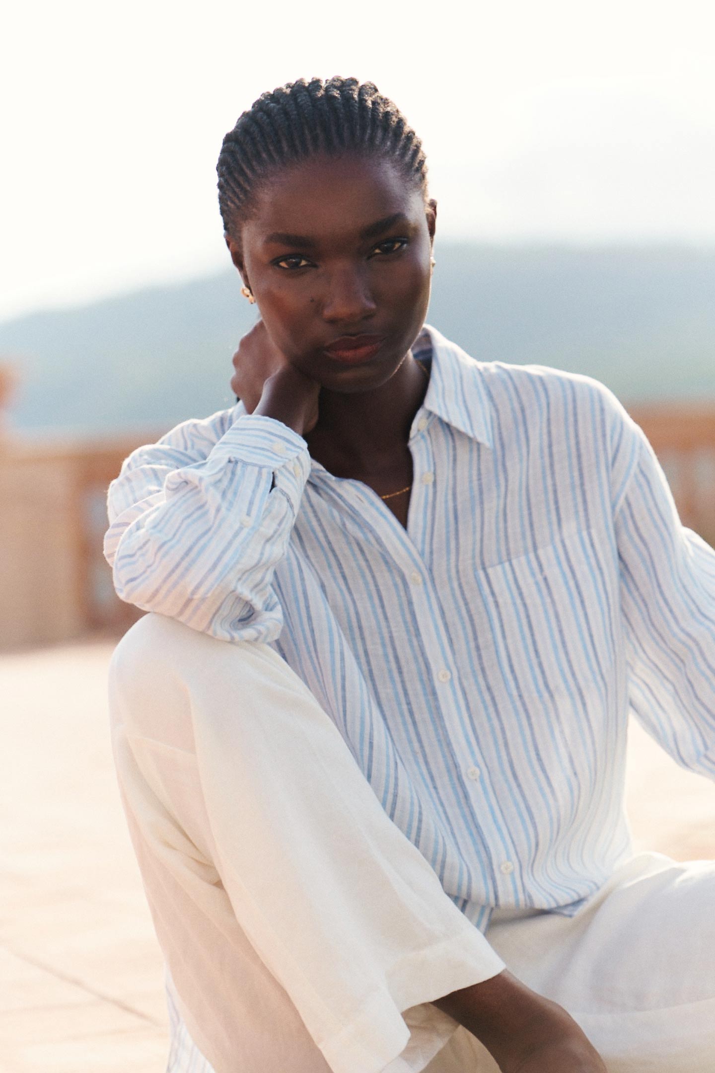 Woman wearing a Gant striped button-down shirt and light trousers, seated outdoors in soft natural light.