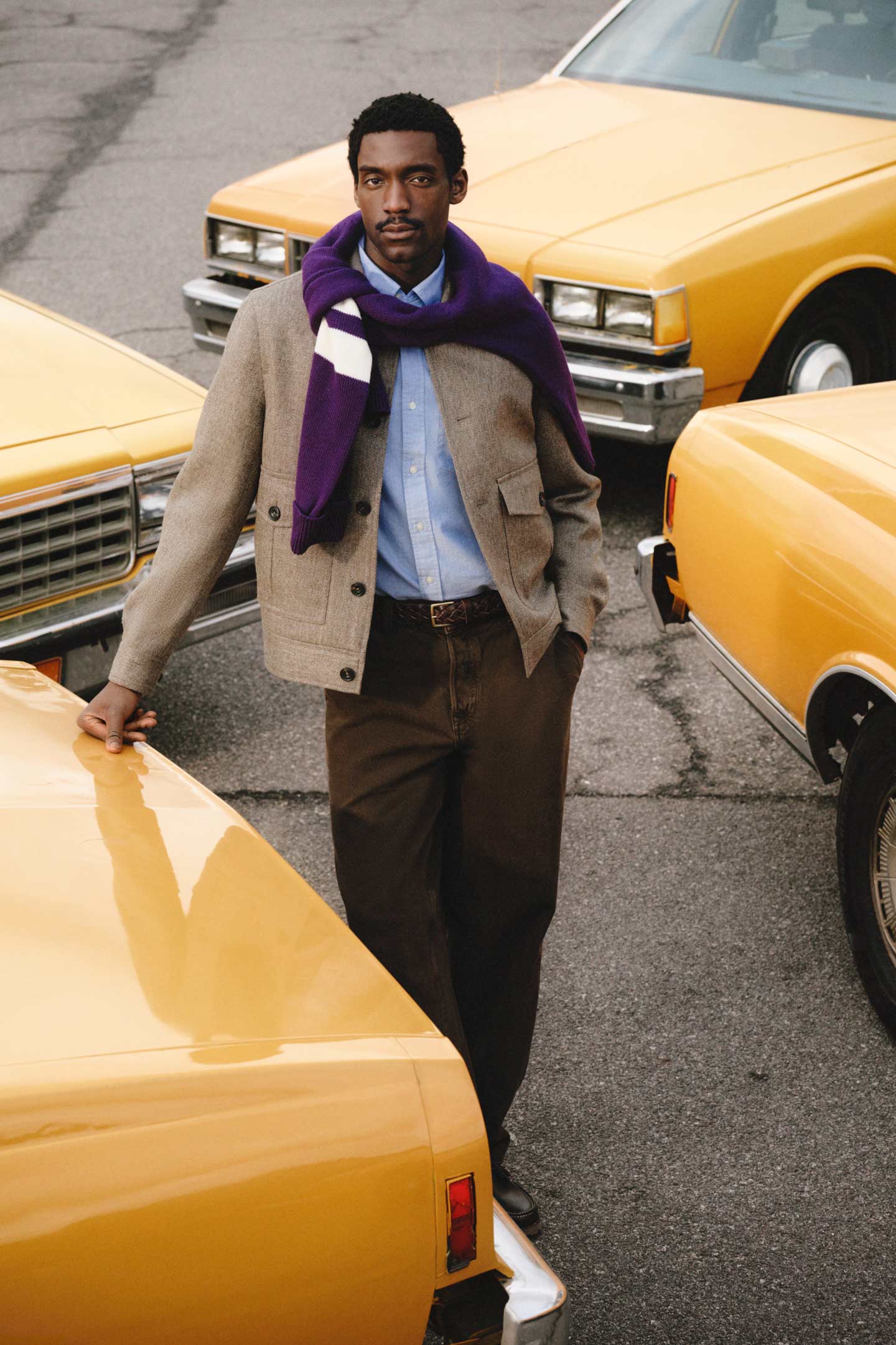 Model styled in a Gant wool jacket over a button-down shirt, pants and scarf, standing between yellow taxis.